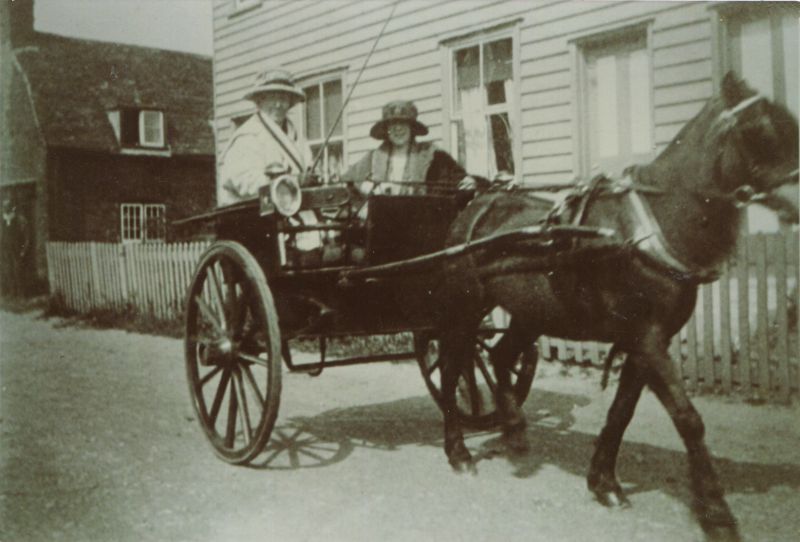  Mrs Josie Condon [ Mary Josephine Condon ] driving a pony and trap at the top of The Lane. The passenger is Mrs Rhona Keenlyside, her daughter. Josie Mary Condon lived in Vine Cottage up to about 1926. The weatherboard cottages on the right are Smithfield Cottages in Firs Chase. The pony's name was Judy.

Used in 2015 exhibition. 
Cat1 Transport - buses and carriers