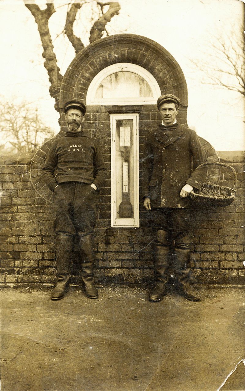  1924 'Billy Oak' Hewes and Titus Mussett. Billy was Harry Mussett's great grandfather and was named 'Oak' after the smack. 

The picture shows the barometer in the wall by The Square on Coast Road - the wall and case are still there, but the barometer has gone. Titus Mussett is holding an oyster tendel.

The name on Bill Oak Hewes' jersey is NANCY, RHYC. 
Cat1 Mersea-->Coast Road Cat2 Families-->Mussett Cat3 Families-->Hewes