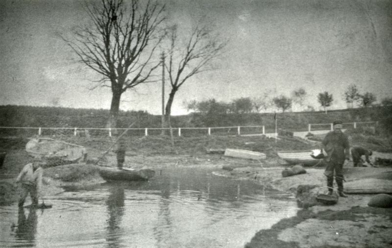  Hove Creek, with Coast Road behind. Oysterman on the right with a tendle on his arm and another on the ground, wearing the canvas smock much favoured by many of the waterfront men.

Now nearly silted up. Clarke Mussett standing, Jack Mole washing winkles [Owen Fletcher].

Before 1928 as there are no houses built by the side of Hove Hill. [RG] 
Cat1 Mersea-->Creeks, fleets, channels, saltings Cat2 Mersea-->Coast Road Cat3 People-->Fishermen and Seamen